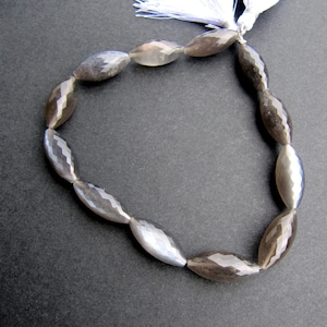 May include: A strand of faceted, oval-shaped beads in shades of grey and silver. The beads are strung together, forming a loop. A white tassel is attached to the top of the strand. The beads are approximately 2.5 cm in length.