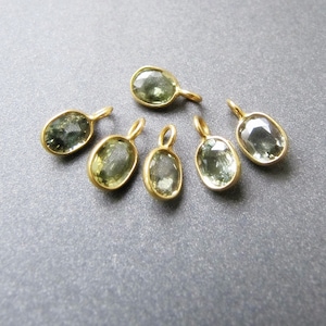 May include: Six oval-shaped green gemstone pendants with gold settings and jump rings. The pendants are arranged in a semi-circle on a grey surface.