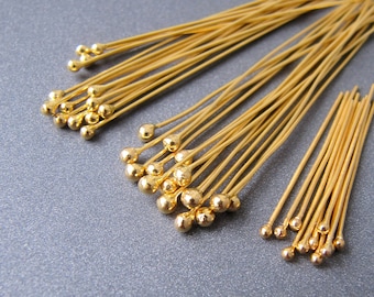 May include: A close-up of a group of gold-colored head pins with round heads. The pins are arranged in a fan shape, with the heads facing the viewer. The pins are about 1 inch long.