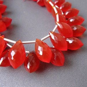 May include: A strand of bright red gemstone beads, each bead is faceted and shaped like a teardrop. The beads are strung on a silver wire.