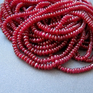 May include: A strand of deep red ruby beads, each bead is round and slightly elongated, creating a textured look. The beads are piled together in a loose heap.