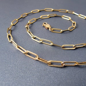 May include: A gold chain necklace with a delicate, flat, oval link design. The chain is laid out in a loose coil on a gray surface.