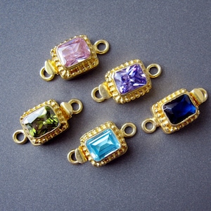 May include: Five gold-toned connector charms with different colored gemstones. Each charm features a square-shaped gemstone with a gold-toned frame and a loop on each side. The gemstones are pink, purple, green, blue, and dark blue.