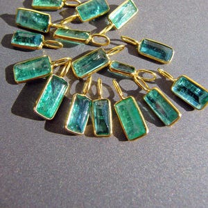 May include: A collection of rectangular green gemstone pendants, each encased in a thin gold-toned metal bezel setting with a small attached jump ring loop at the top. The stones exhibit varying shades of translucent teal and emerald green hues.