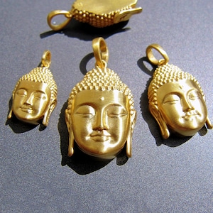 May include: Gold-colored Buddha head pendants with closed eyes and detailed hair. The pendants are in various sizes, with a larger pendant at the top and three smaller ones below. The pendants are on a gray surface.