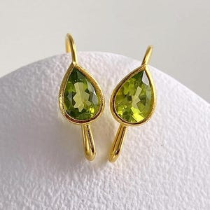 Puede incluir: Un par de pendientes colgantes de oro con piedras preciosas de peridoto facetadas en forma de pera. Cada pendiente tiene un engaste de oro amarillo liso que enmarca la piedra verde vibrante, suspendido de un gancho de alambre de oro.