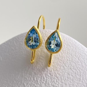 May include: A pair of drop earrings featuring faceted, pear-shaped blue topaz gemstones set in textured, matte yellow gold bezels. The earrings are suspended from simple gold wire hooks, showcasing a clear, bright blue hue and a polished finish.