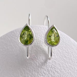 Può includere: Un paio di orecchini pendenti con gemme di peridoto verde lime sfaccettate a forma di pera, incastonate in metallo color argento. Ogni orecchino presenta un semplice gancio curvo per l'attacco, mostrando una tonalità verde brillante e chiara.