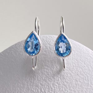 May include: A pair of silver drop earrings featuring faceted, pear-shaped blue gemstones. Each stone is set in a textured silver bezel with a curved silver wire hook for attachment. The design is symmetrical, showcasing clear blue stones.