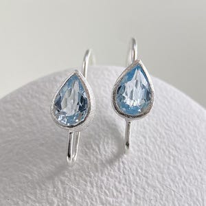 May include: A pair of drop earrings featuring faceted, pear-shaped light blue gemstones set in textured silver metal. Each earring has a curved silver wire hook for attachment, showcasing a clear, pale blue stone with a pointed base.