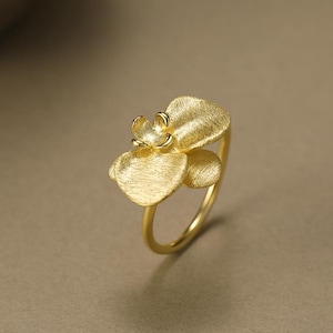 Peut inclure: Une bague dorée avec un motif floral. La bague a un anneau délicat et une fleur texturée avec plusieurs pétales. La fleur est le point central, avec un centre détaillé et des surfaces texturées. La bague est probablement en métal.