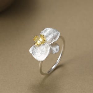 Op de afbeelding: Een zilveren ring met een bloemdesign en een goudkleurig centrum. De bloem heeft getextureerde bloemblaadjes en is gezet op een dunne, zilveren band. De ring is een sieraad.