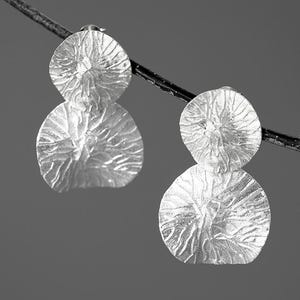 May include: A pair of silver-colored earrings with a textured, layered design. Each earring features a small, round top piece connected to a larger, petal-shaped bottom piece. The earrings are suspended against a gray background.