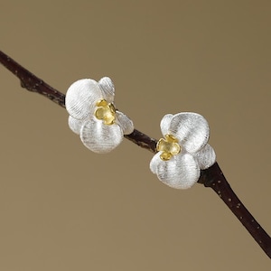 Puede incluir: Pendientes de botón florales plateados con centros dorados. Los pendientes están diseñados para parecer delicadas flores. Se muestran en una rama marrón sobre un fondo neutro.