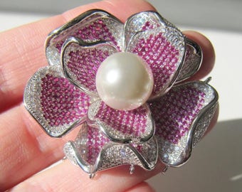 May include: A silver flower brooch with a pearl in the center. The flower is made of small pink and white stones.