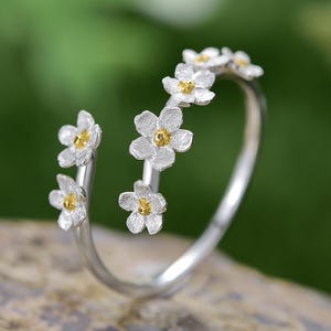 Puede incluir: Un anillo plateado con un diseño floral. El anillo presenta pequeñas flores blancas con centros amarillos. Las flores están dispuestas a lo largo de la banda, creando una estética delicada e inspirada en la naturaleza. El anillo está sobre un fondo verde borroso.