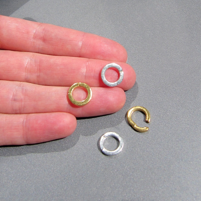May include: Four silver and gold jump rings, a type of jewelry finding, are shown on a hand. The jump rings are open and ready to be used in jewelry making.