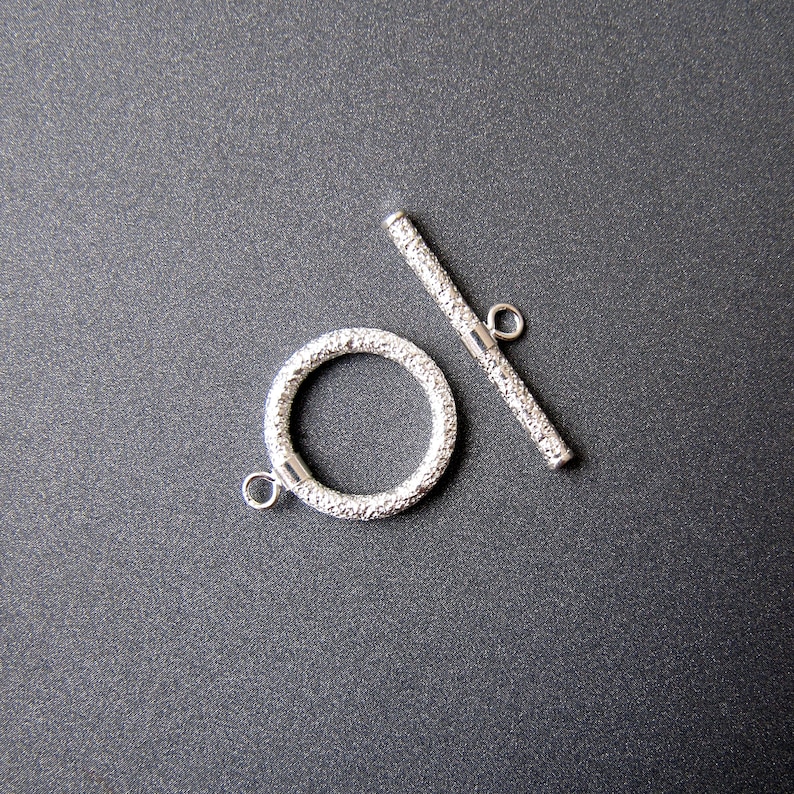 May include: Silver toggle clasp with a textured finish. The clasp has a round ring and a straight bar.