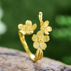 Puede incluir: Un anillo dorado con un diseño floral. El anillo presenta tres flores texturizadas dispuestas en un racimo. La banda se curva elegantemente, y las flores tienen pétalos detallados. El fondo es verde suave, creando un efecto natural.