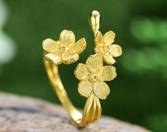 May include: A gold-colored ring featuring a floral design with three textured flowers. The ring's band curves gracefully, and the flowers are arranged in a cluster. The background is a soft green, suggesting a natural setting.