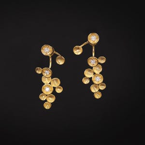 Peut inclure: Une paire de boucles d'oreilles dorées au design unique. Chaque boucle d'oreille présente une série d'éléments circulaires texturés de tailles variables, reliés par de fines tiges. De petites pierres précieuses scintillantes sont serties au centre de certains cercles. Le fond est noir.