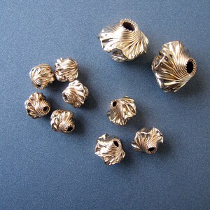 K&ouml;nnte beinhalten: Eine Sammlung von neun goldfarbenen Metallperlen mit einem wirbelnden, strukturierten Design. Die Perlen sind auf einer blauen Oberfl&auml;che angeordnet.