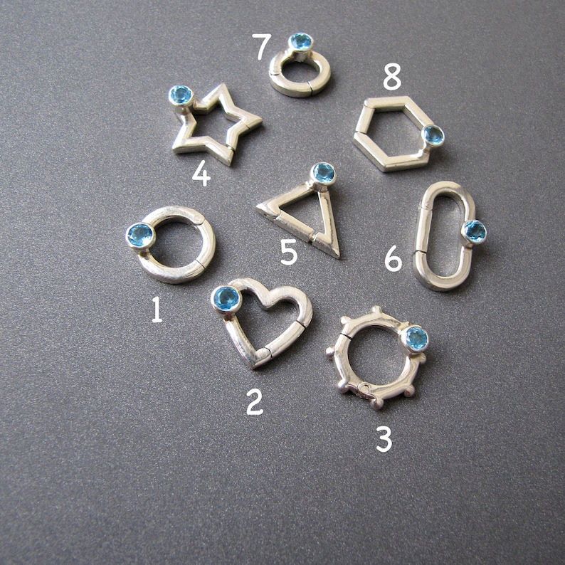 May include: Eight silver charms with blue gemstones. Each charm is a different shape: circle, heart, star, triangle, hexagon, and three more circles with different designs.