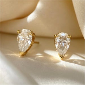 Può includere: Un paio di orecchini a lobo con pietre trasparenti sfaccettate a forma di pera, incastonate in metallo color oro giallo lucido. Ogni orecchino è fissato con una montatura a tre griffe che mantiene saldamente in posizione la gemma a goccia.