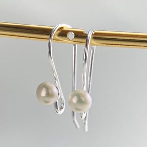 May include: A pair of silver earrings featuring a curved wire design with a single round white pearl attached to the end of each piece. The earrings are suspended from a thin gold-toned horizontal bar, showcasing a polished metallic finish.