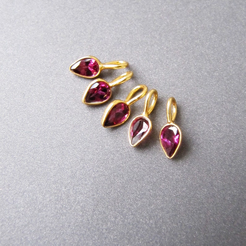 Op de afbeelding: Vijf gouden traanvormige bedels met roze edelstenen. De bedels hebben een lus aan de bovenkant om aan sieraden te bevestigen.