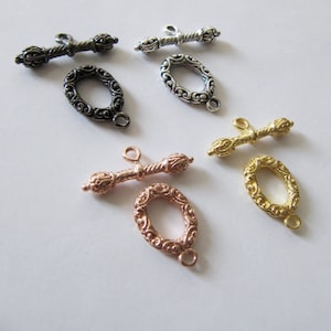 Pode incluir: Quatro fechos de encaixe ornamentados em diferentes acabamentos: prata, ouro, ouro rosa e latão antigo. Cada fecho apresenta um laço oval decorativo e uma barra torcida com extremidades ornamentadas.
