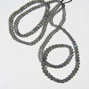 May include: A strand of gray labradorite beads with iridescent blue and green flashes. The beads are strung on a black cord and are approximately 6mm in diameter.