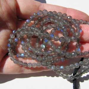 May include: A strand of gray labradorite gemstone beads with iridescent blue flashes. The beads are round and faceted, and the strand is strung on a black cord.