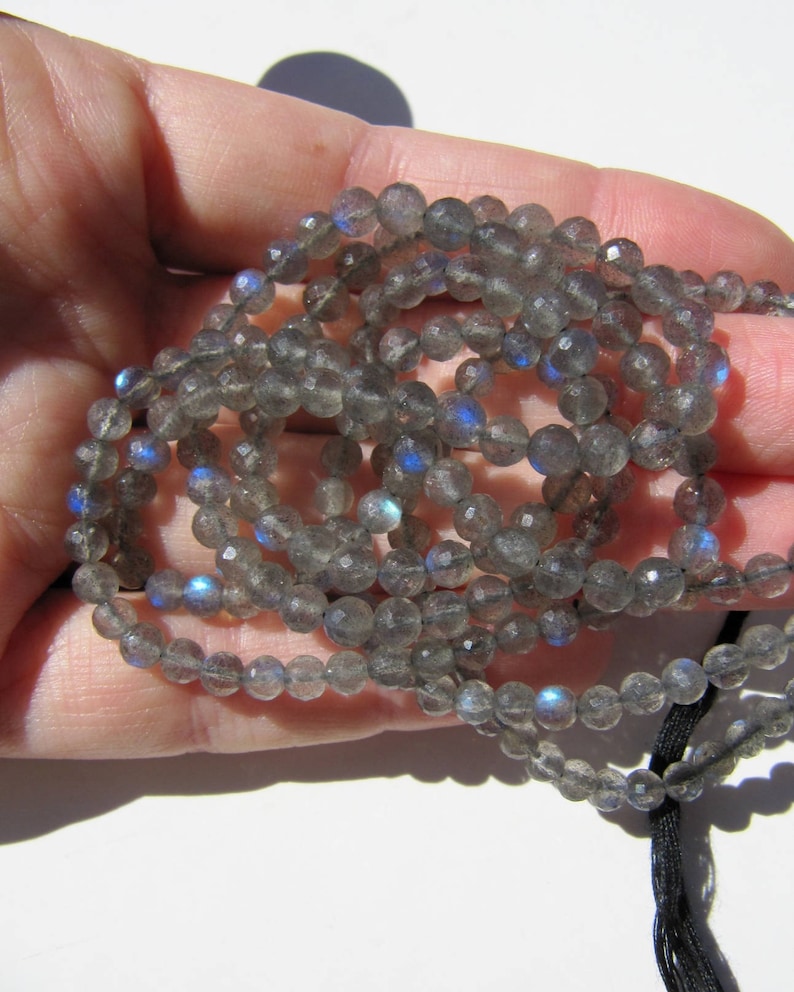 May include: A strand of gray labradorite gemstone beads with iridescent blue flashes. The beads are round and faceted, and the strand is strung on a black cord.
