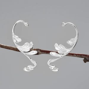 Peut inclure: Une paire de boucles d'oreilles argentées avec un motif délicat de feuilles. Les boucles d'oreilles sont faites d'un métal fin et flexible et sont conçues pour s'enrouler autour de l'oreille.