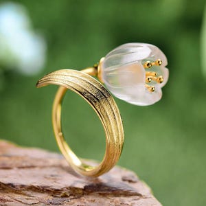 Op de afbeelding: Een goudkleurige ring met een delicaat bloemdesign. De bloem heeft een doorschijnende, bloembladachtige structuur met kleine, goudkleurige accenten. De ringband heeft een getextureerde, bladachtige uitstraling, wat bijdraagt aan de natuurlijke esthetiek. De achtergrond is groen.