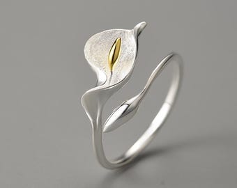 May include: A silver ring designed as a calla lily. The flower features a white petal and a gold-coloured stamen. The ring has a smooth, polished finish and wraps around the finger. This is a floral-themed piece of jewellery.