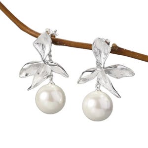 May include: A pair of silver-toned earrings featuring a floral design with three petals and a large, round, white pearl. The earrings are hanging from a thin, brown branch against a white background.