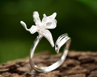 May include: A silver-colored ring featuring a delicate, open flower design. The ring's band curves around, with a leaf-like detail on one side. The flower has detailed petals, creating a nature-inspired jewelry piece.