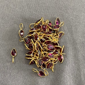 Op de afbeelding: Een stapel gouden hangerbedels met paarse edelsteenaccenten. De bedels zijn traanvormig en hebben een lus aan de bovenkant om aan een ketting of armband te bevestigen.
