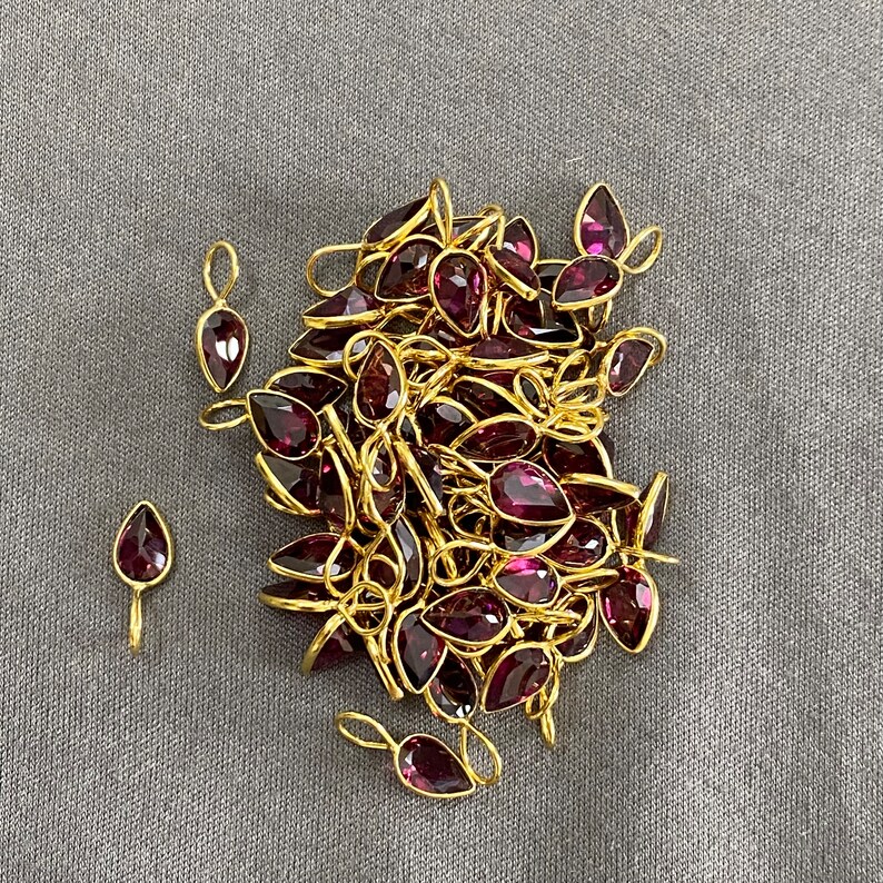 Op de afbeelding: Een stapel gouden hangerbedels met paarse edelsteenaccenten. De bedels zijn traanvormig en hebben een lus aan de bovenkant om aan een ketting of armband te bevestigen.