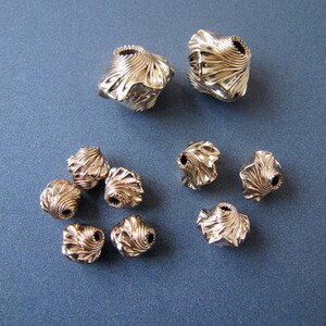 K&ouml;nnte beinhalten: Eine Sammlung von zehn goldfarbenen Metallperlen mit einem einzigartigen, strukturierten und leicht gekr&auml;uselten Design. Die Perlen sind in einem Halbkreis auf einer blauen Oberfl&auml;che angeordnet.