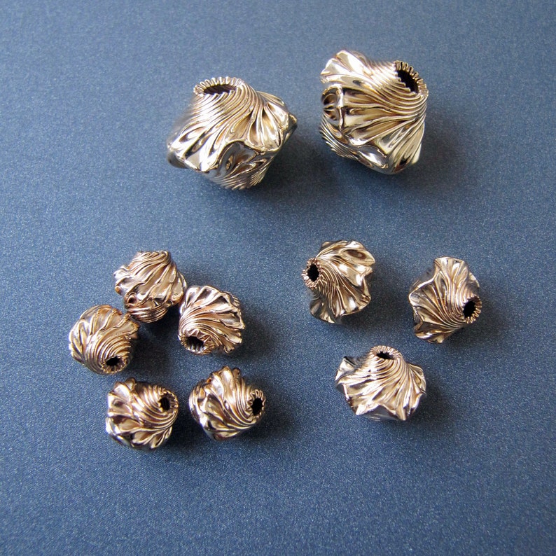 K&ouml;nnte beinhalten: Eine Sammlung von zehn goldfarbenen Metallperlen mit einem einzigartigen, strukturierten und leicht gekr&auml;uselten Design. Die Perlen sind in einem Halbkreis auf einer blauen Oberfl&auml;che angeordnet.