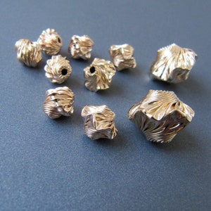 K&ouml;nnte beinhalten: Eine Sammlung von zehn goldfarbenen Metallperlen mit komplizierten, strukturierten Designs. Die Perlen sind in verschiedenen Formen und Gr&ouml;&szlig;en, wobei einige eine abgerundete Form und andere eine eckigere Form haben.