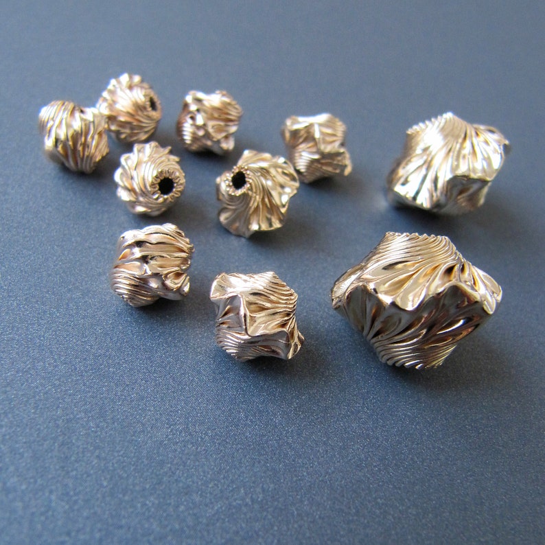 K&ouml;nnte beinhalten: Eine Sammlung von zehn goldfarbenen Metallperlen mit komplizierten, strukturierten Designs. Die Perlen sind in verschiedenen Formen und Gr&ouml;&szlig;en, wobei einige eine abgerundete Form und andere eine eckigere Form haben.
