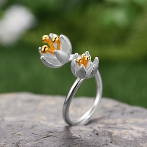 Peut inclure: Une bague en argent avec deux motifs de fleurs blanches. Les fleurs ont des centres dorés.