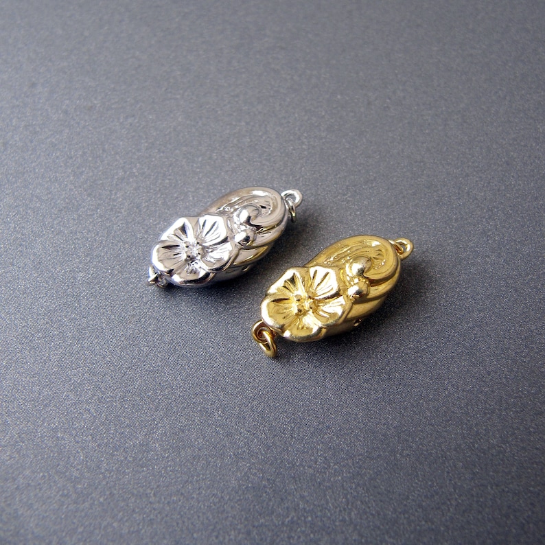 May include: Two ornate metal clasps, one silver and one gold, shaped like a flower with a rounded oval base.