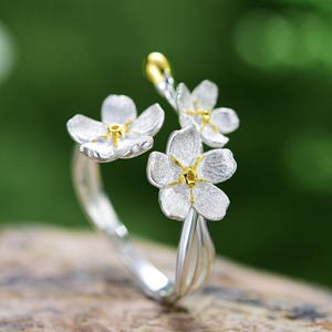 Könnte beinhalten: Silberring mit einem zarten Design aus drei weißen Blüten mit goldenen Zentren. Die Blüten sind auf einem Stiel angeordnet, der sich um den Finger wickelt.