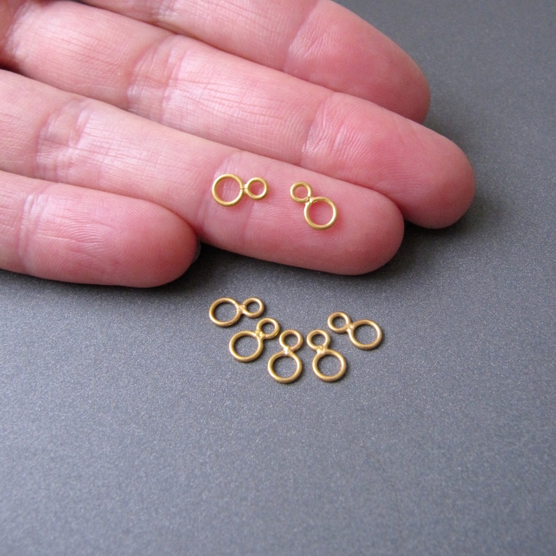 May include: Gold-toned jump rings, also known as double loop rings, are shown on a hand. The rings are small and delicate, perfect for jewelry making.