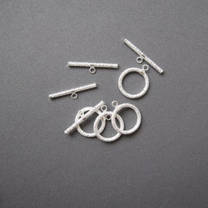 May include: Silver toggle clasps with a textured finish. The clasps are in the shape of a bar and a circle. There are five clasps in total.